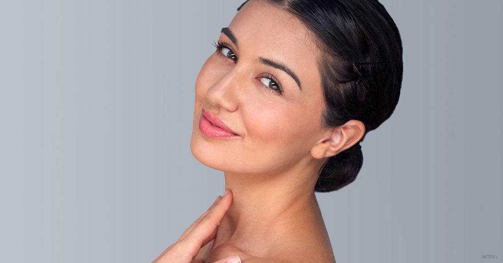 Woman with dark hair in a low bun smiling with her hand resting on her neck (model)